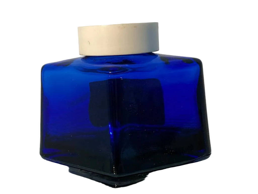 A cobalt blue glass bottle with a square shape and a white screw cap, resting on a reflective glass surface against a white background.
