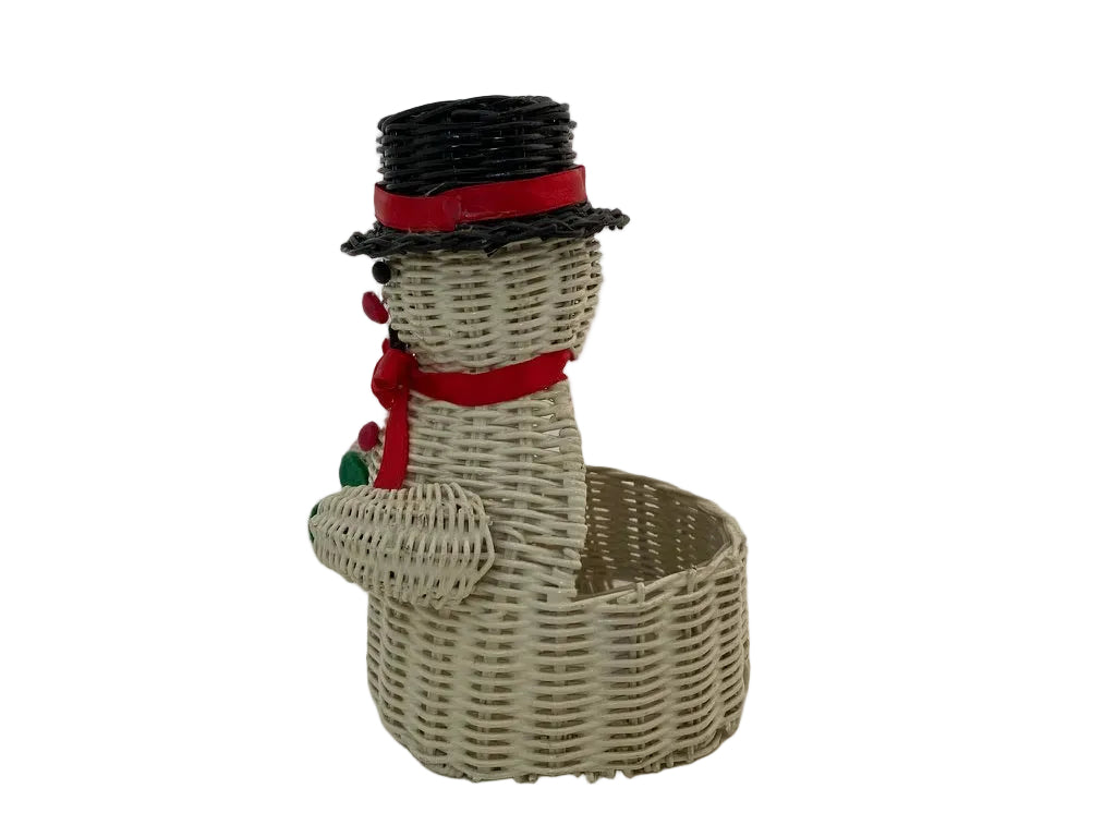 A decorative snowman figurine made from woven natural wicker, featuring a black top hat with a red band, a red scarf, and small red and green pom-poms, standing beside an open round basket base on a plain white background.