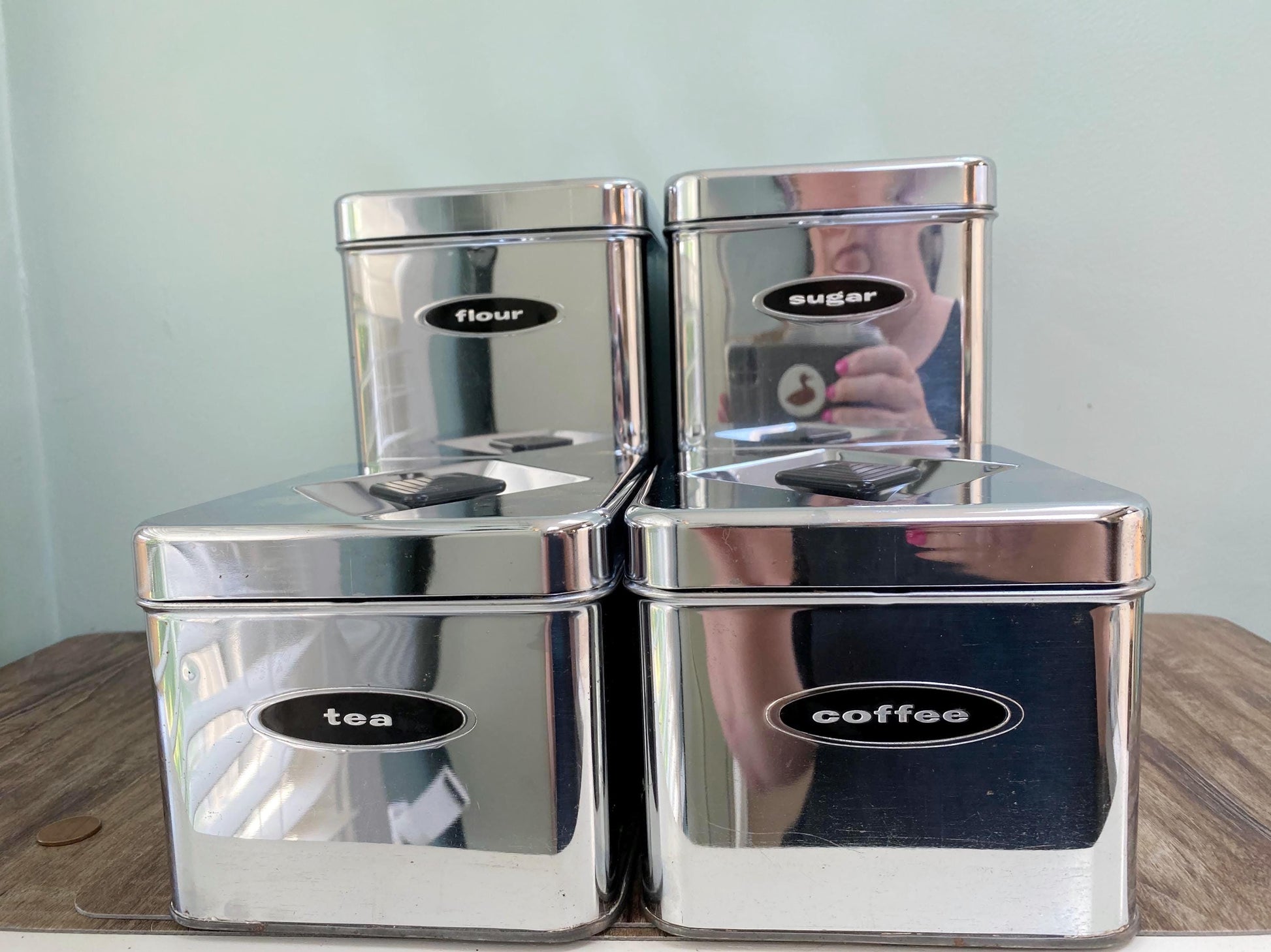 Four reflective stainless steel canisters for flour, sugar, tea, and coffee, arranged in a stack on a wooden surface against a pale green wall.