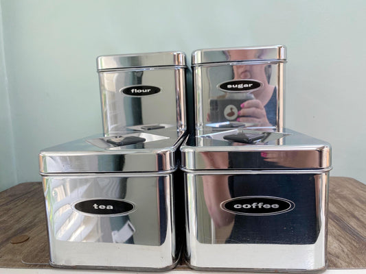 Four reflective stainless steel canisters for flour, sugar, tea, and coffee, arranged in a stack on a wooden surface against a pale green wall.